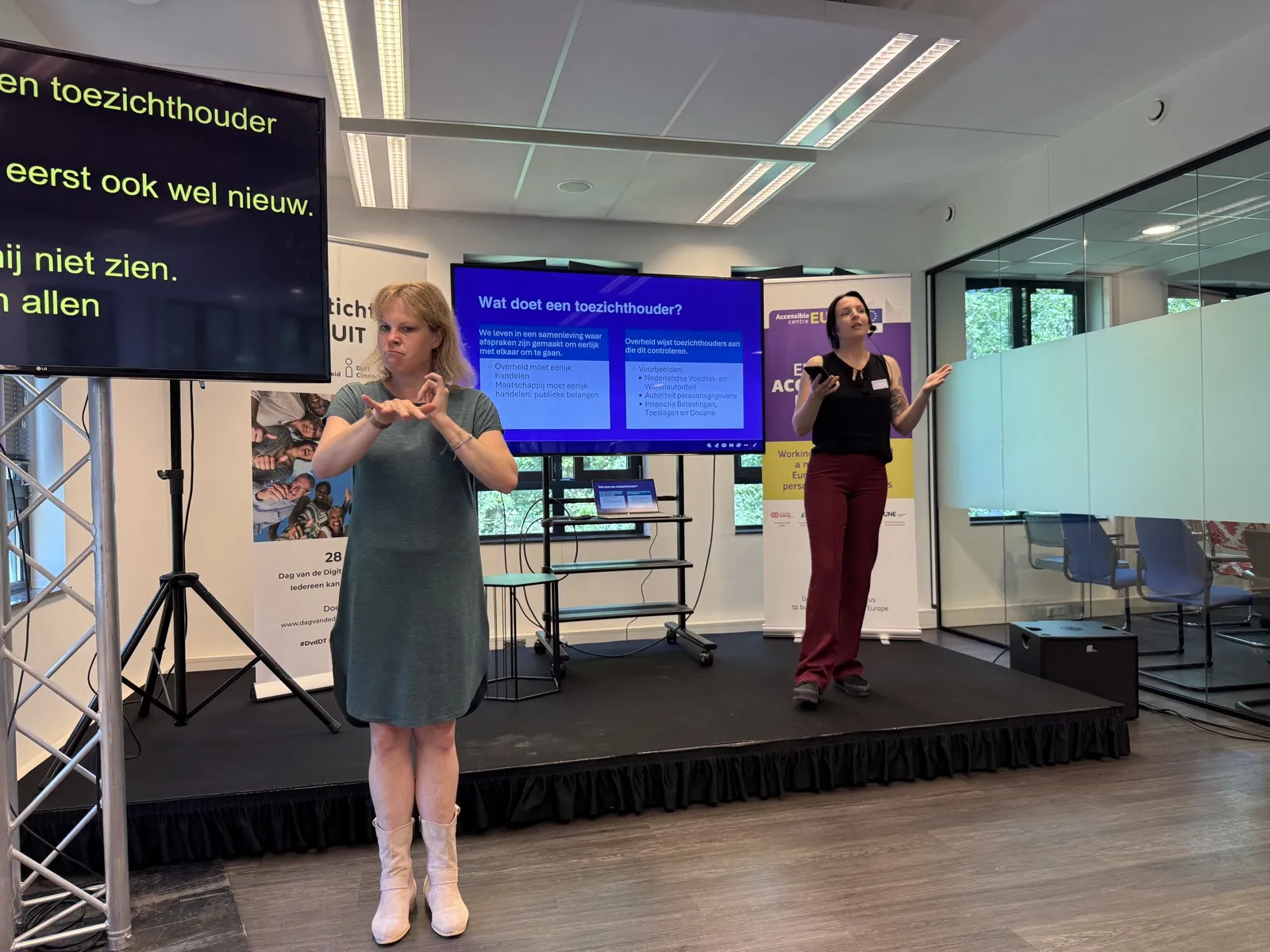 Caitlin de Rooij van de ACM op het podium tijdens de Dag van Digitale Toegankelijkheid, met een gebarentolk naast haar en een slide met de tekst Wat doet een toezichthouder.